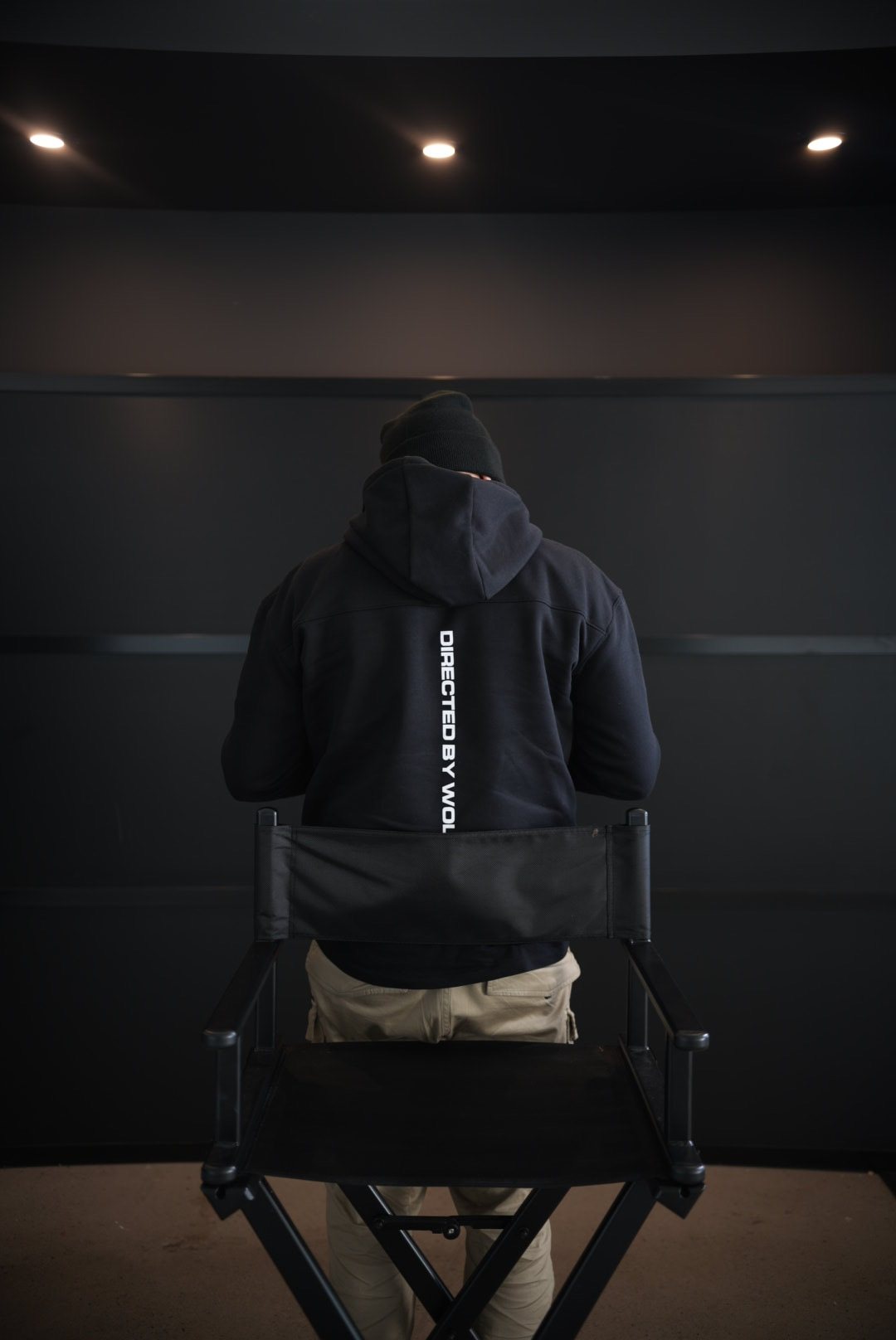 Wolf from behind in a director's chair, DIRECTED BY WOLF on the back of his hoodie